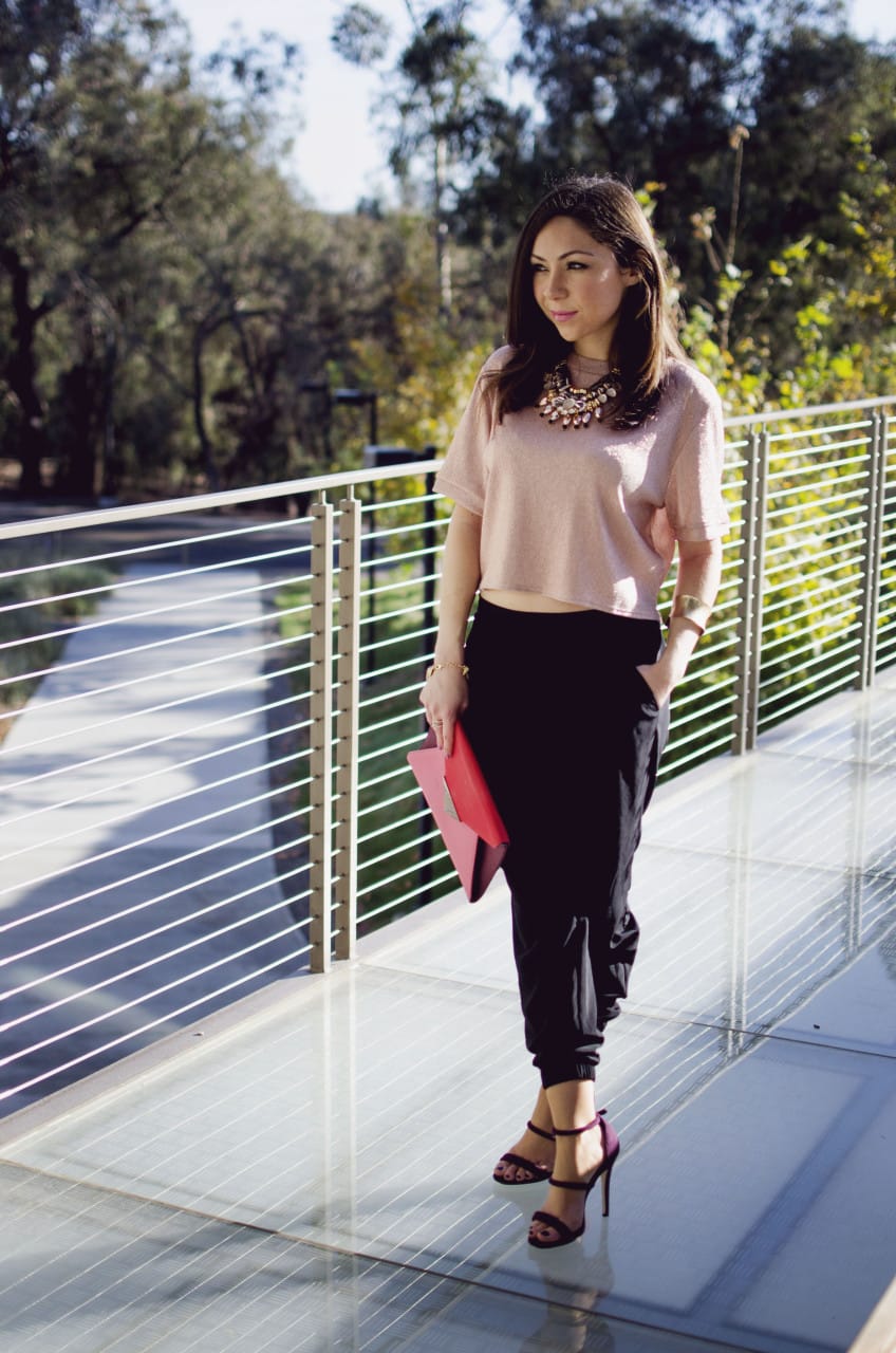elegant holiday party look - Blogger Nihan standing and showing her Holiday Party Outfit; a pink metallic top, a big statement necklace, black jogger pants, maroon highly and a color block clutch.