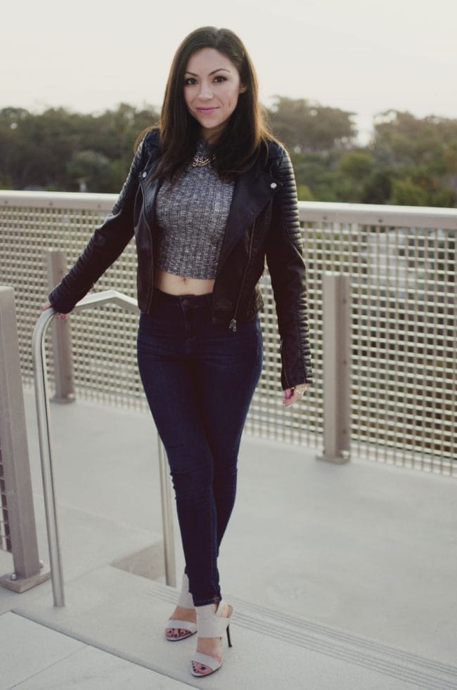 Blogger Nihan smiling and posing to show her edgy cool and sexy outfit; a gray top, blue jeans, leather jacket, neutral high-heels and a red and pink clutch.