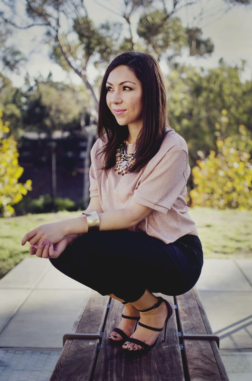 Blogger Nihan sitting and showing her Holiday Party Outfit; a pink metallic top, a big statement necklace, black jogger pants, maroon high heels and trendy jewelry.