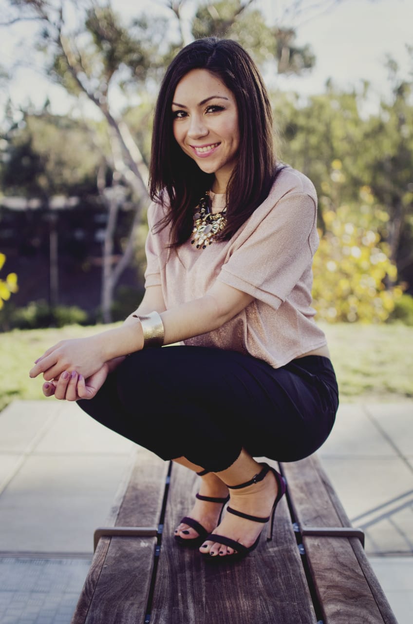 elegant holiday party look - Blogger Nihan sitting and showing her Holiday Party Outfit; a pink metallic top, a big statement necklace, black jogger pants, maroon high heels and trendy jewelry.