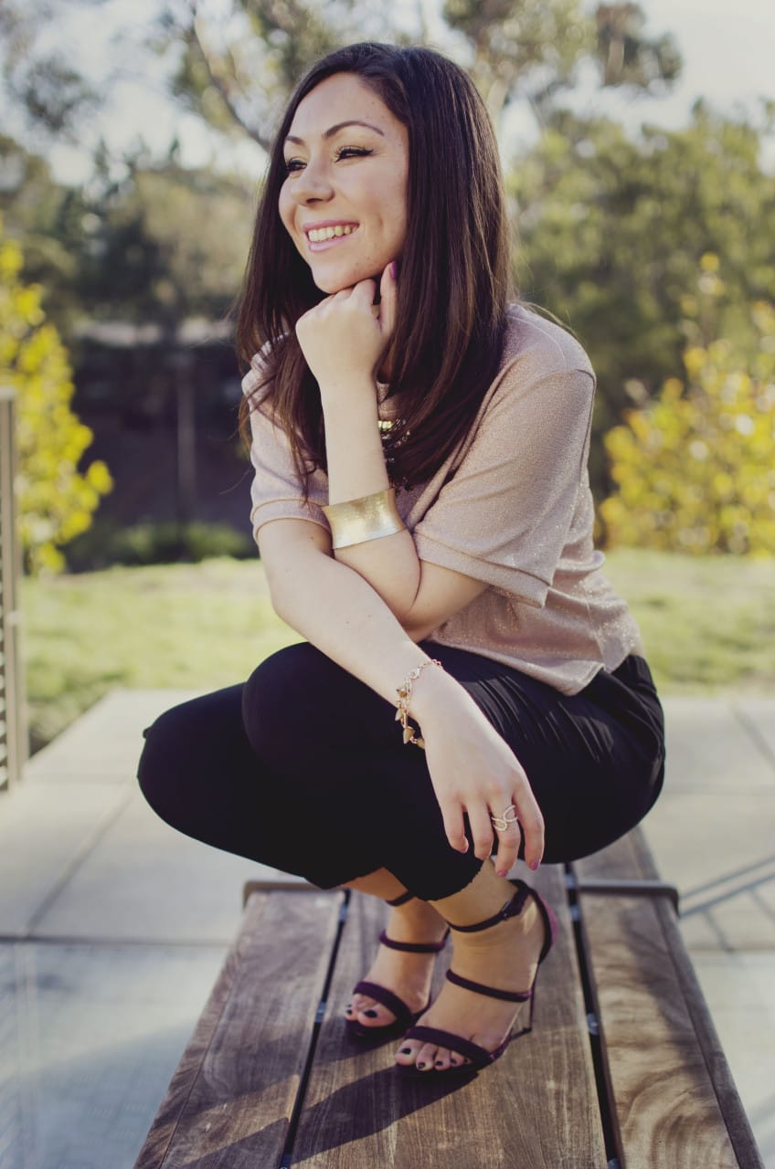 Blogger Nihan sitting and showing her Holiday Party Outfit; a pink metallic top, a big statement necklace, black jogger pants, maroon high heels and trendy jewelry.