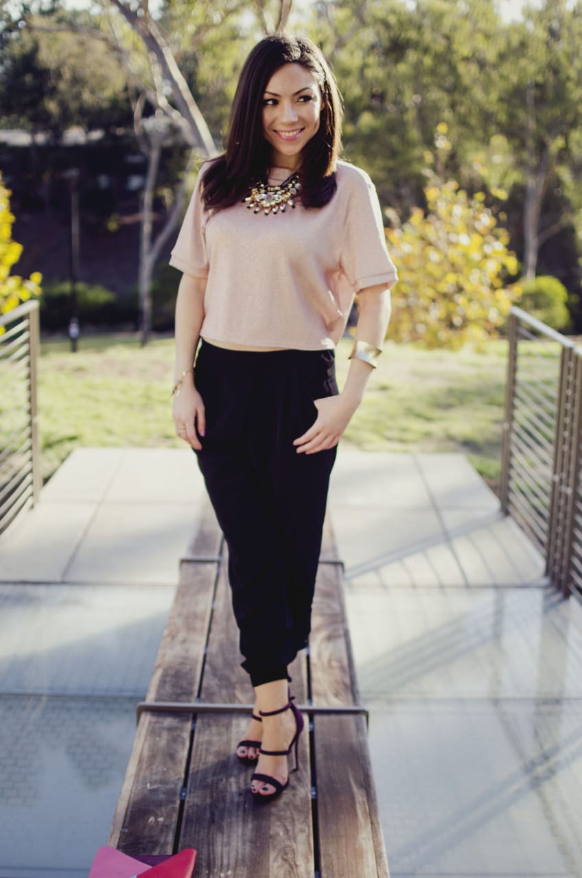 Blogger Nihan standing and showing her Holiday Party Outfit; a pink metallic top, a big statement necklace, black jogger pants, maroon highly and a color block clutch.