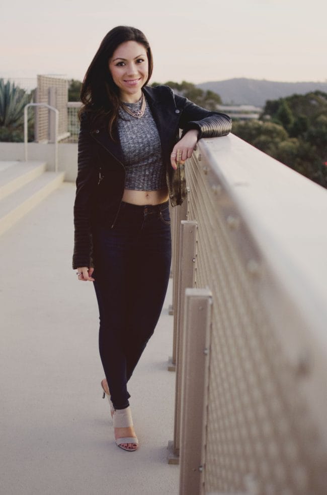 Blogger Nihan smiling and posing to show her edgy cool and sexy outfit; a gray top, blue jeans, leather jacket, neutral high-heels and a red and pink clutch.