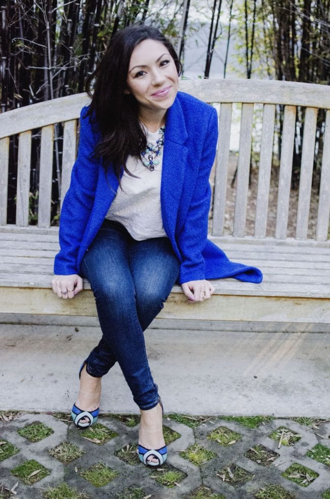 Fashion model smiling and showing her outfit featuring a blue oversized coat, white top, floral green and blue necklace and and Topshop jeans while sitting on a bench