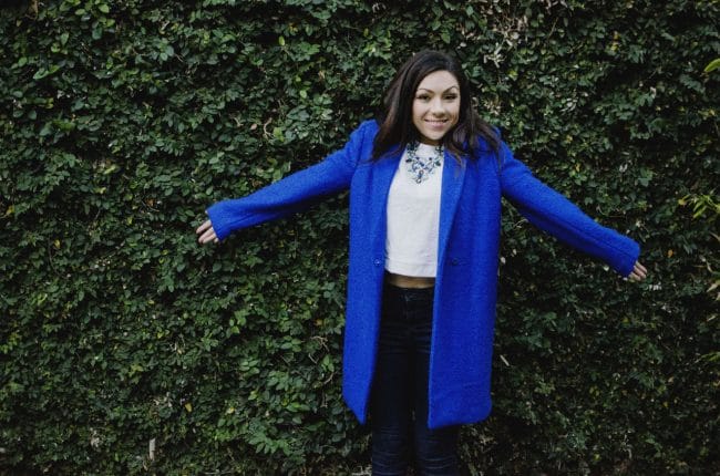 Fashion model being goofy and funny while showing her outfit featuring a blue oversized coat, white top, floral green and blue necklace and and Topshop jeans